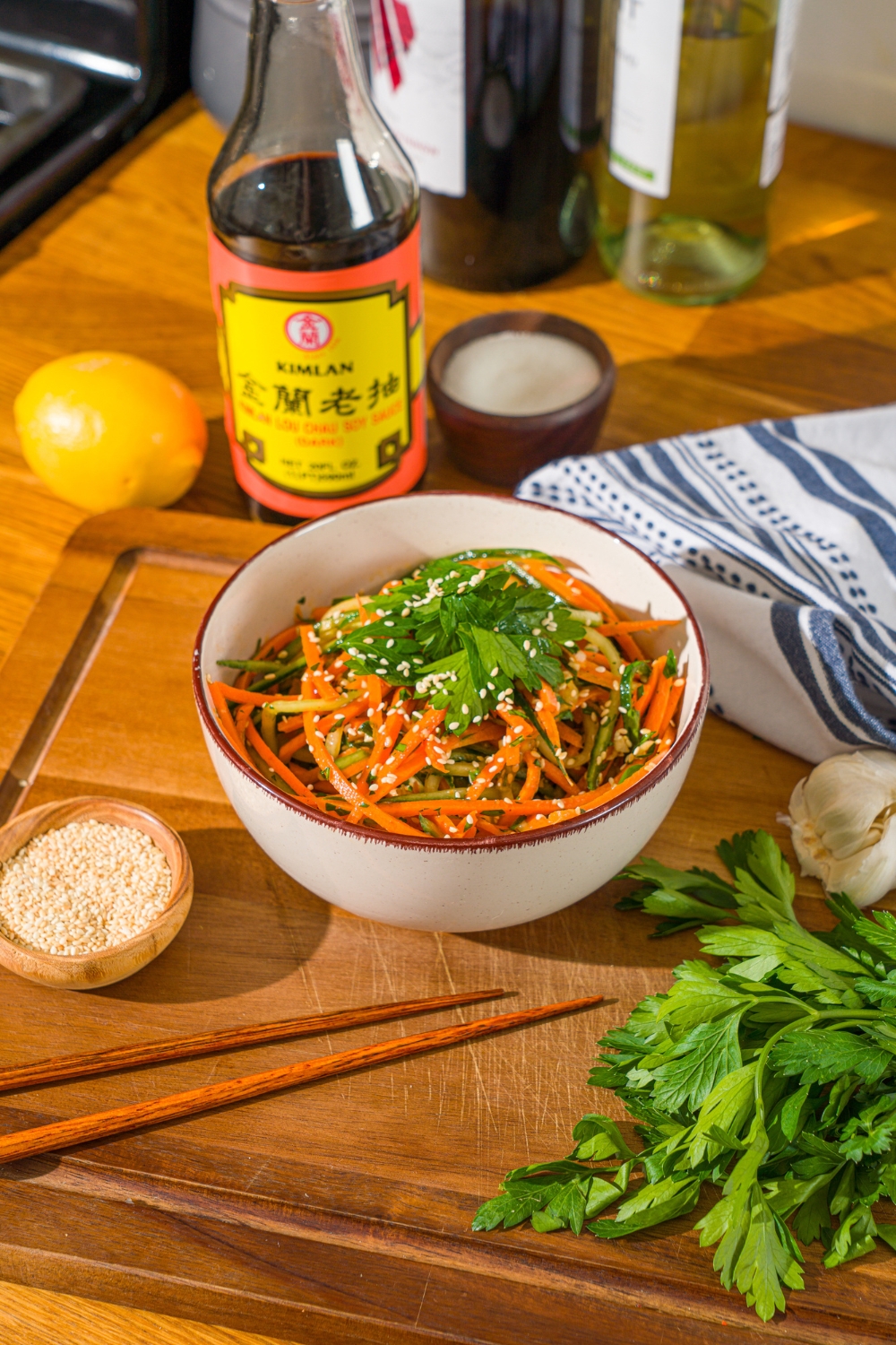 A white bowl with carrot cucumber salad tossed in dressing and garnished with fresh parsley and sesame seeds. The bowl is on a wooden board with a pair of chopsticks and small bowl of sesame seeds.