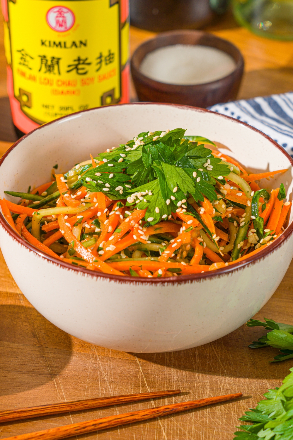 A white bowl with carrot cucumber salad tossed in dressing and garnished with fresh parsley and sesame seeds. The bowl is on a wooden board with a pair of chopsticks and a jar of soy sauce.