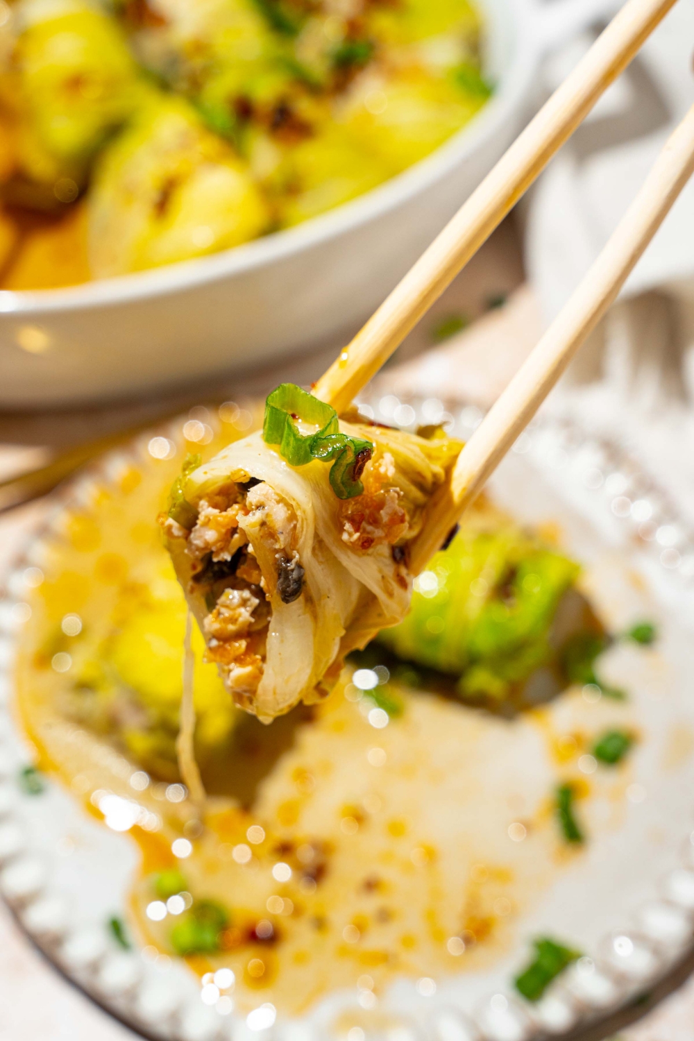 A close up of a pair of chopsticks holding a cabbage dumpling topped with chili oil, sesame seeds, and sliced green onion with a bite removed. There is a plate of dumplings blurred in the background.