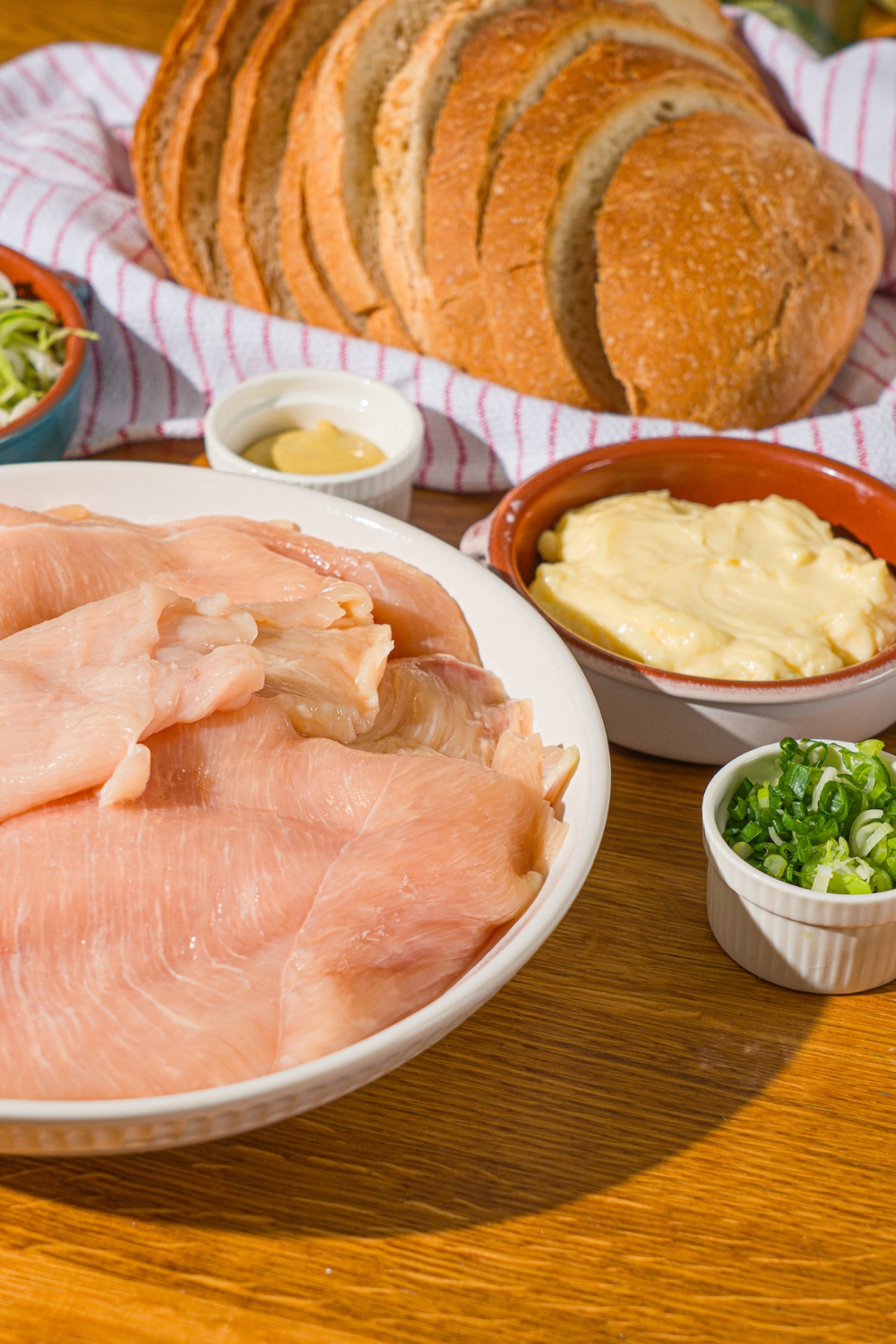 A wooden counter with several bowls containing ingredients to make a chicken melt including chicken, bread, mayo, sliced green onions, mustard, and more.
