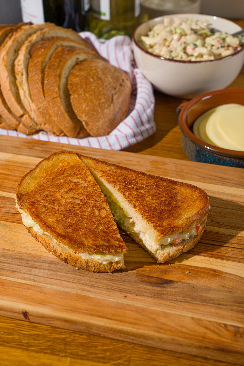 A wooden board with a chicken melt sandwich sliced in half. The board is on a wooden counter with a basket of bread and bowl of chicken.