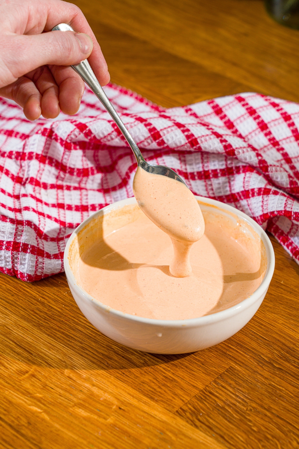 A white bowl with comeback sauce with a spoon dipping into the bowl. The bowl is on a wooden counter with a red checkered napkin.