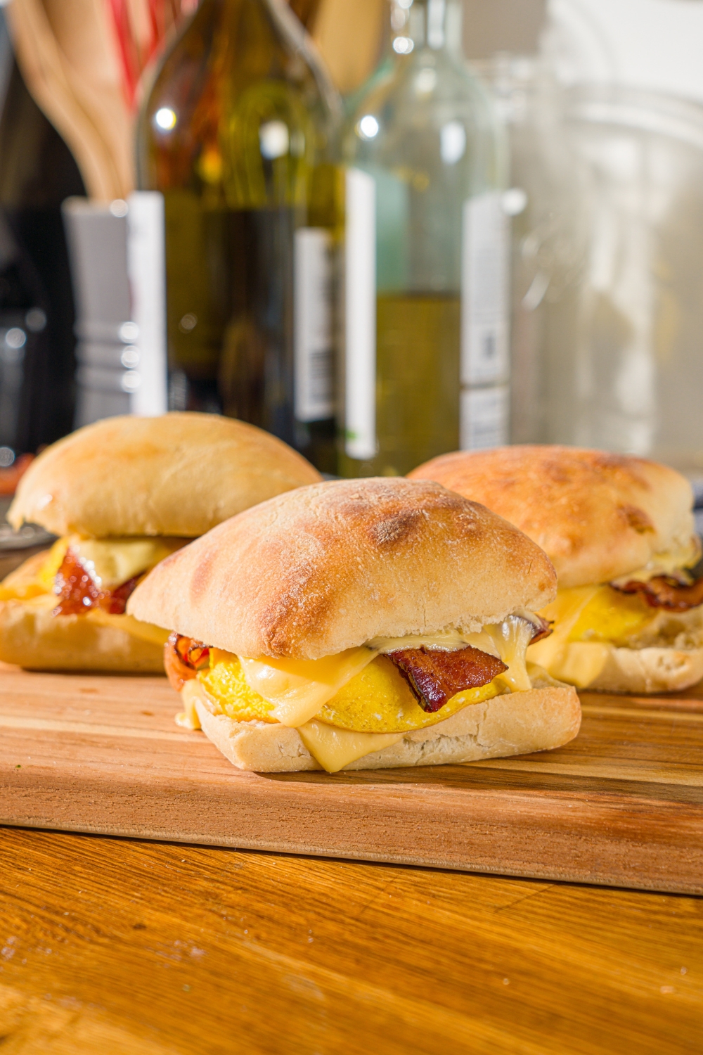 A wooden cutting board with cooked Starbucks gouda bacon egg sandwiches. The board is on a wooden counter.