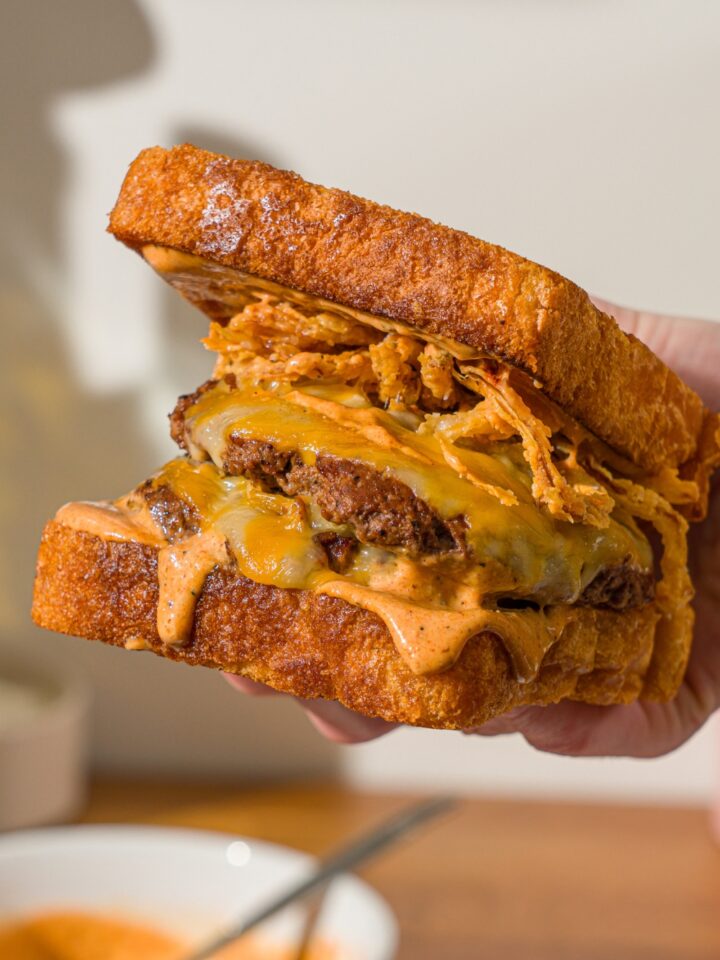 A hand holding a copycat Whataburger patty melt. There is a bowl of Whataburger patty melt sauce on a wooden counter blurred in the background.