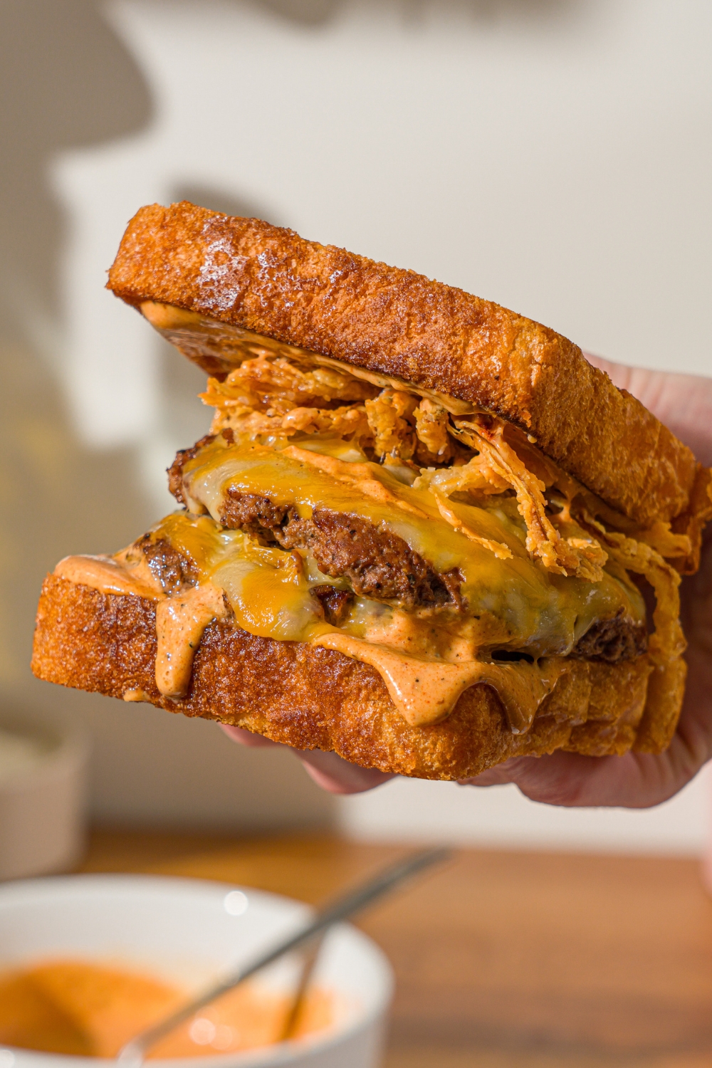 A hand holding a copycat Whataburger patty melt. There is a bowl of Whataburger patty melt sauce on a wooden counter blurred in the background.