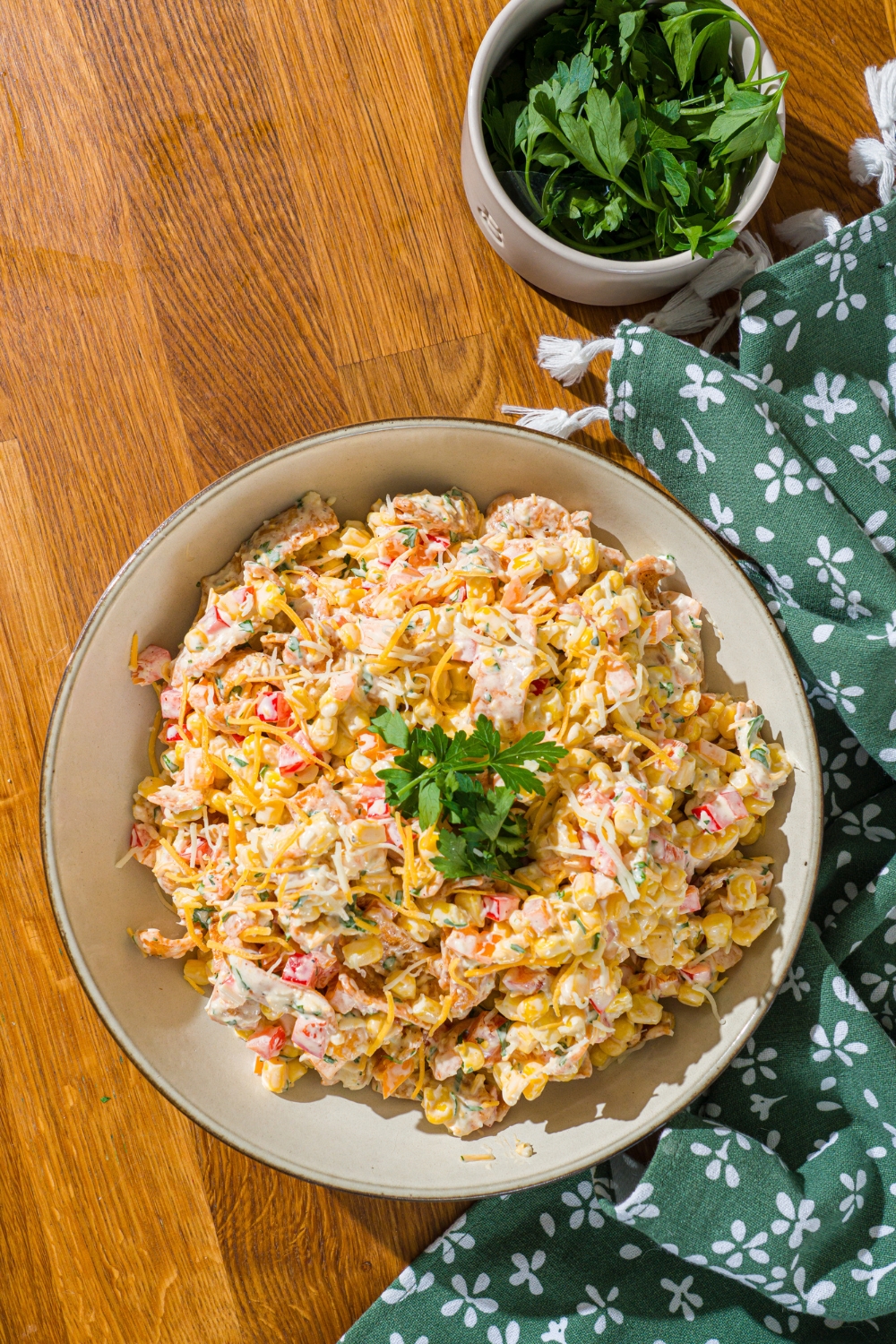 A white bowl with corn salad with chili cheese fritos garnished with fresh parsley. The bowl is on a wooden counter with a green floral napkin and a bowl of garnishes.