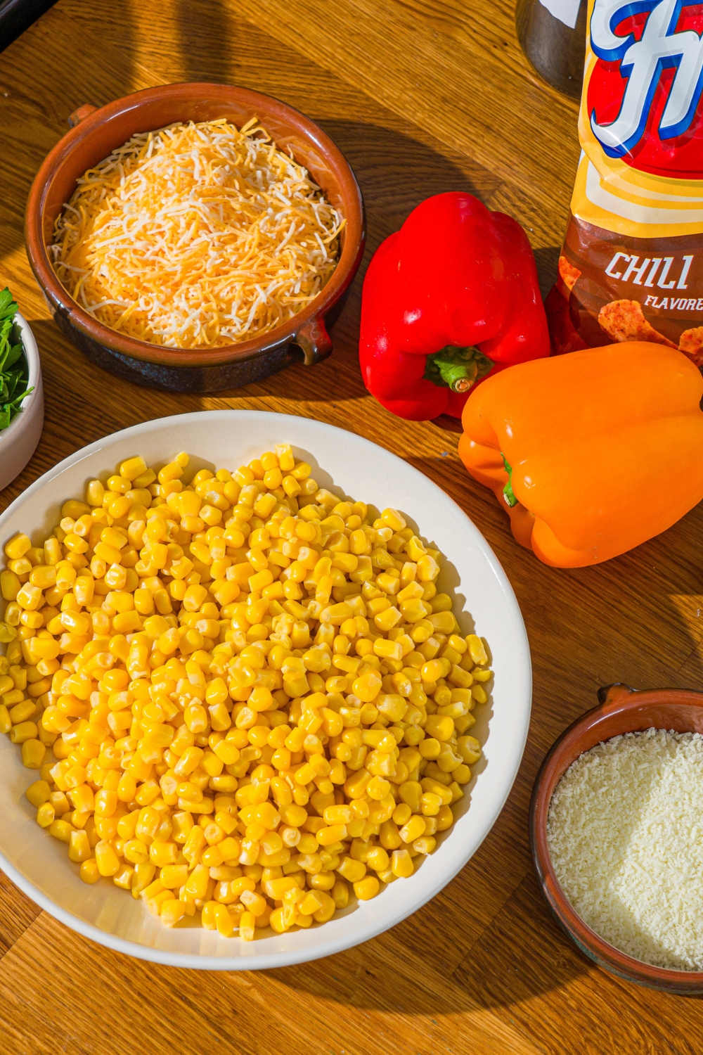 A wooden counter with ingredients to make corn salad with chili cheese fritos including corn, red and yellow bell pepper, shredded cheese, and chili cheese fritos.