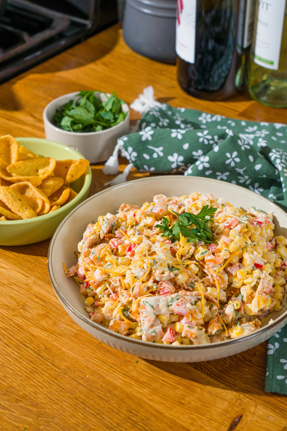 A white bowl with corn salad with chili cheese fritos garnished with fresh parsley. The bowl is on a wooden counter with a green floral napkin and a bowl of garnishes.