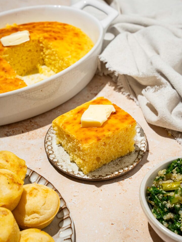 A plate with a slice of cornbread without eggs topped with a pat of butter. The plate is on a tan counter with a baking dish of cornbread and white cloth napkin.