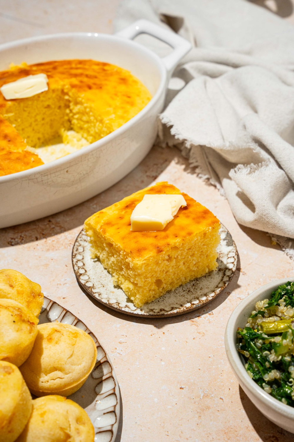 A plate with a slice of cornbread without eggs topped with a pat of butter. The plate is on a tan counter with a baking dish of cornbread and white cloth napkin.