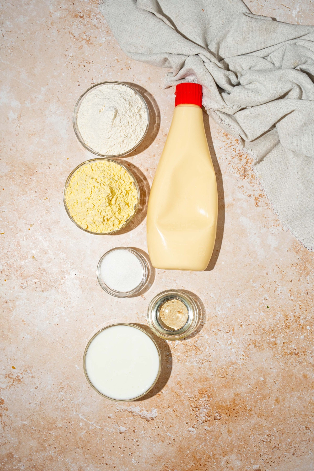A tan counter with bowls containing ingredients to make cornbread without eggs including buttermilk, mayo, sugar, flour, cornmeal, and oil.