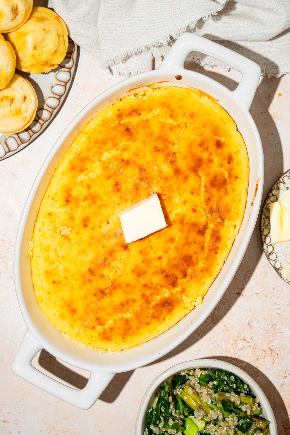 An oval baking dish with baked cornbread without eggs topped with a pat of butter. The dish is on a tan counter with a plate of muffins and white cloth napkin.