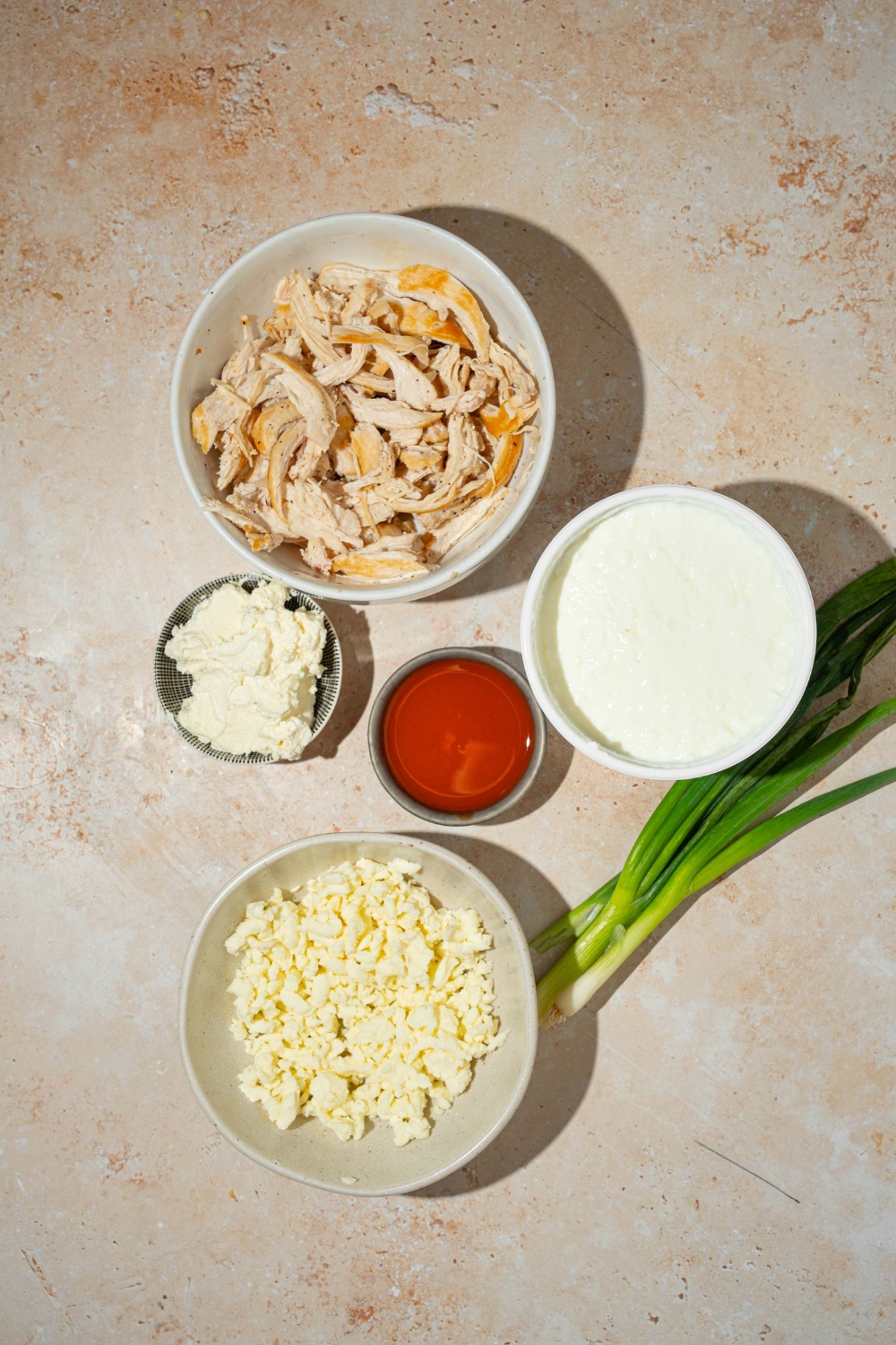 A tan counter with several bowls containing ingredients to make cottage cheese Buffalo chicken dip including shredded chicken, cottage cheese, Greek yogurt, shredded cheese, hot sauce, and green onion.