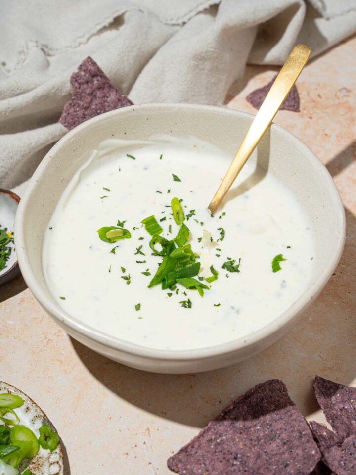 A white bowl with cottage cheese ranch dip garnished with sliced green onion and fresh dill. There is a spoon in the bowl. The bowl is on a tan counter with blue corn tortilla chips.