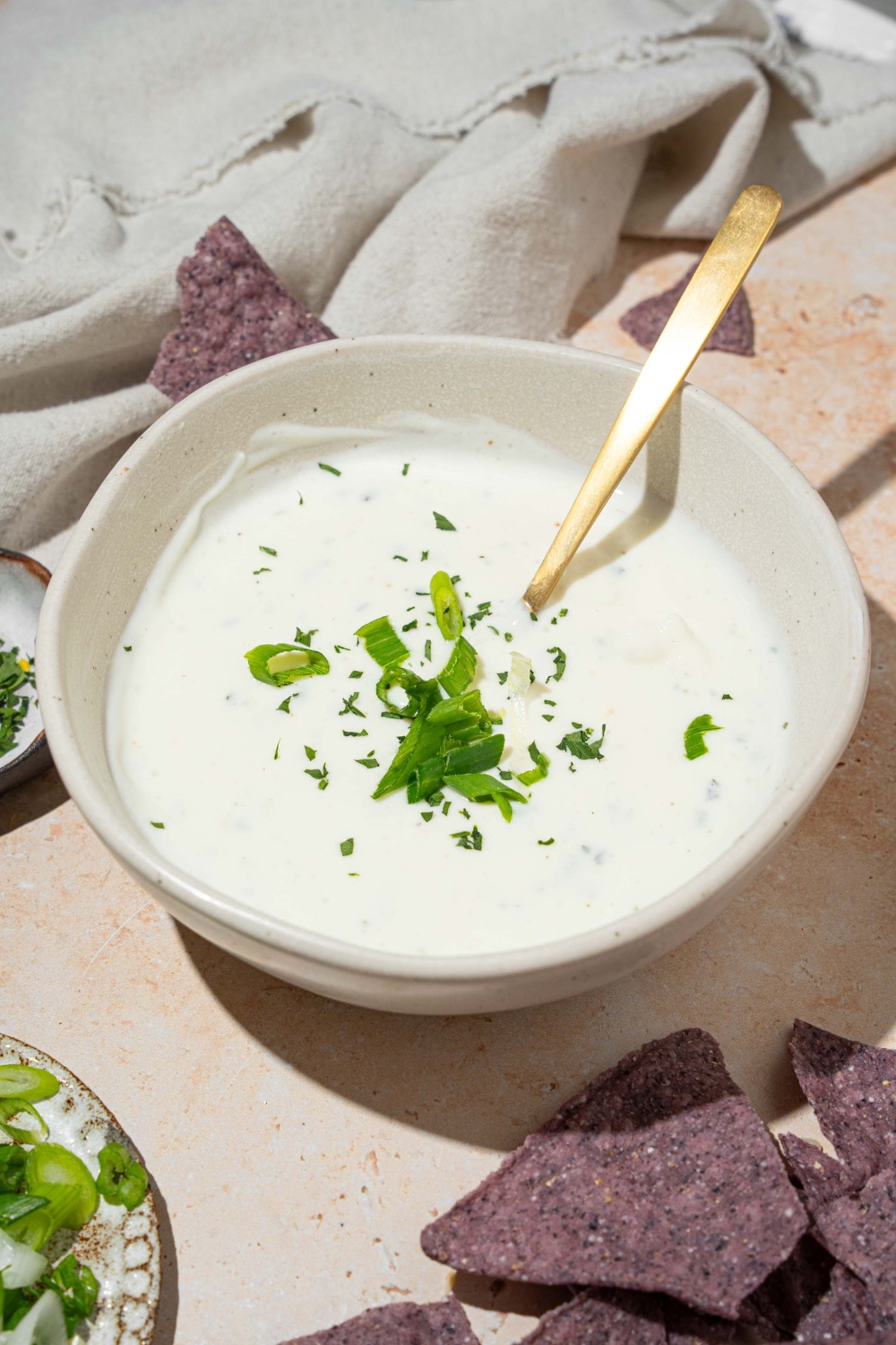 A white bowl with cottage cheese ranch dip garnished with sliced green onion and fresh dill. There is a spoon in the bowl. The bowl is on a tan counter with blue corn tortilla chips.