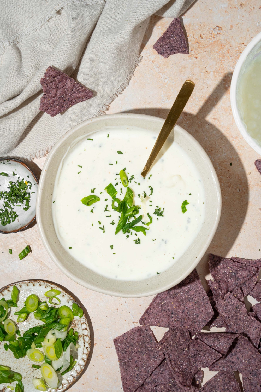 A white bowl with cottage cheese ranch dip garnished with sliced green onion and fresh dill. There is a spoon in the bowl. The bowl is on a tan counter with blue corn tortilla chips.
