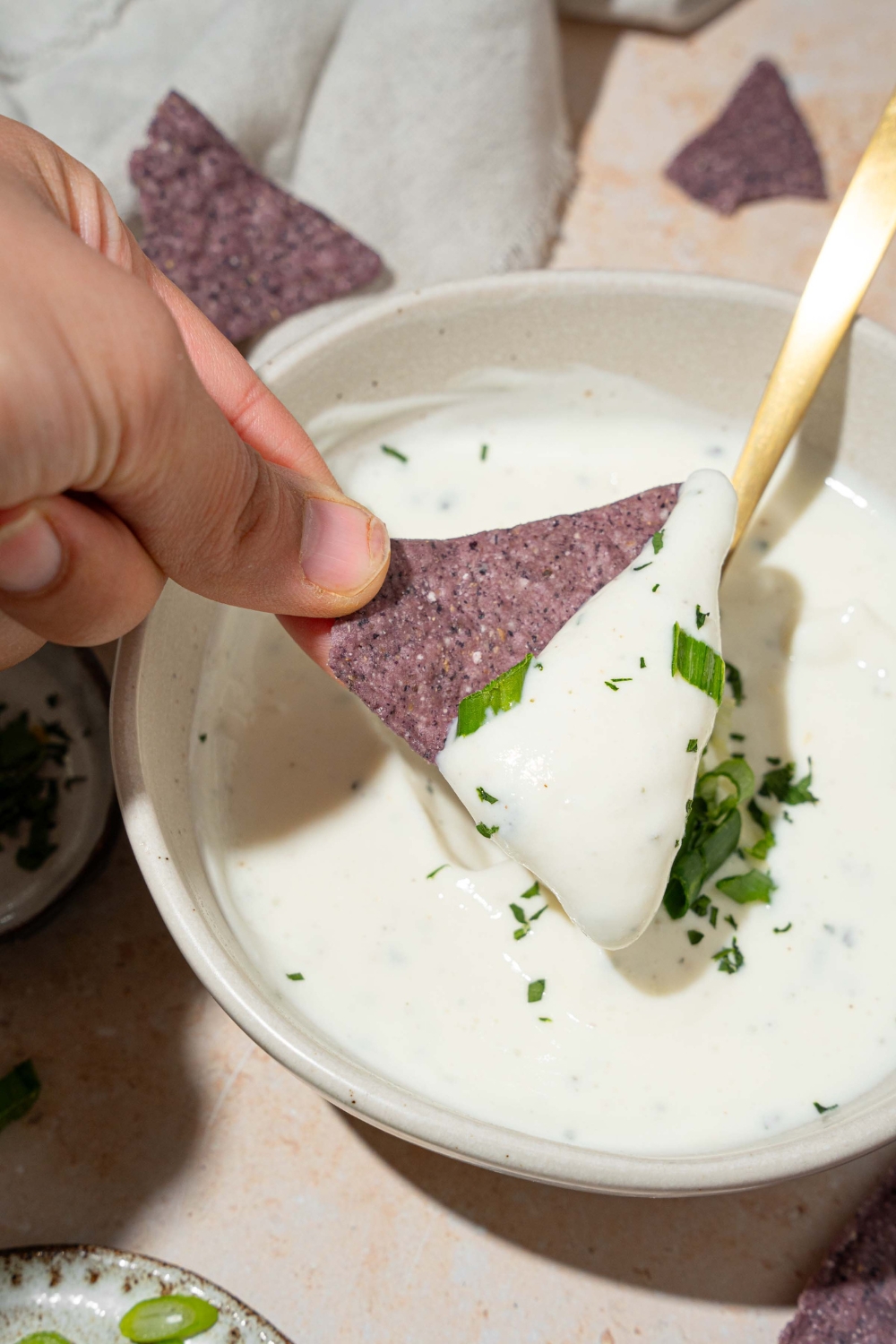 A white bowl with cottage cheese ranch dip garnished with sliced green onion and fresh dill. There is a spoon in the bowl. A hand is dipping a blue corn tortilla chip into the bowl.
