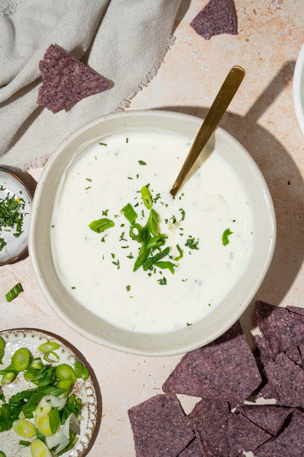 A white bowl with cottage cheese ranch dip garnished with sliced green onion and fresh dill. There is a spoon in the bowl. The bowl is on a tan counter with blue corn tortilla chips.