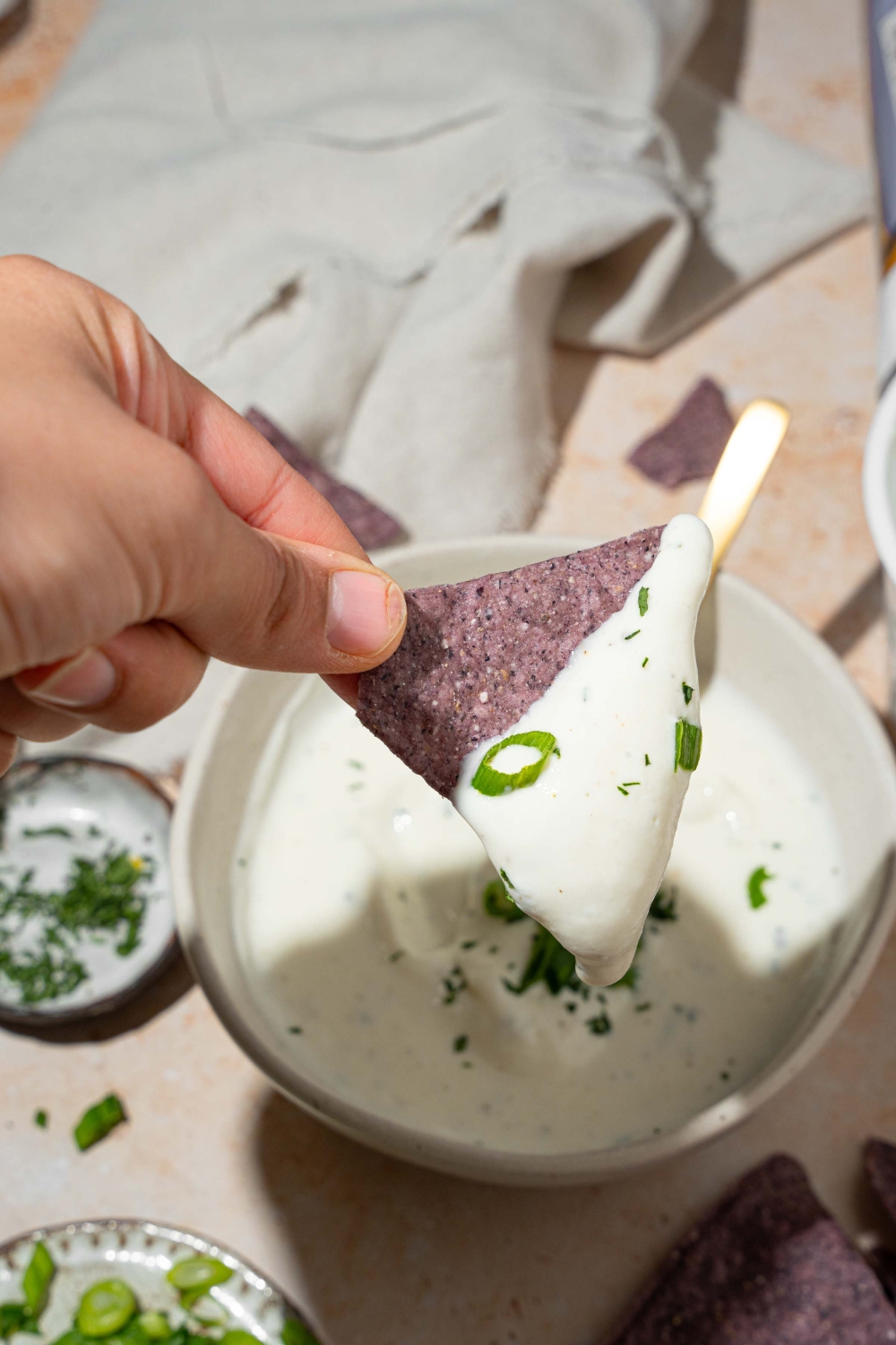 A close up of a hand holding a blue corn tortilla chip dipped in cottage cheese ranch dip. There is a white bowl with cottage cheese ranch dip on a tan counter blurred in the background.