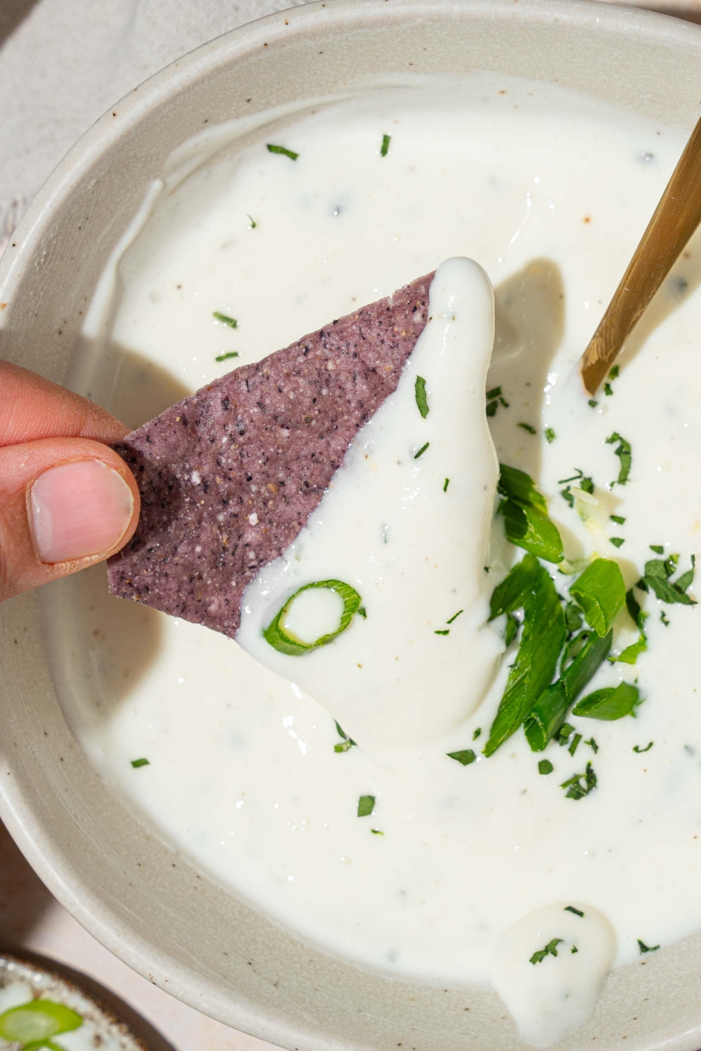 A white bowl with cottage cheese ranch dip garnished with sliced green onion and fresh dill. There is a spoon in the bowl. A hand is dipping a blue corn tortilla chip in the bowl.