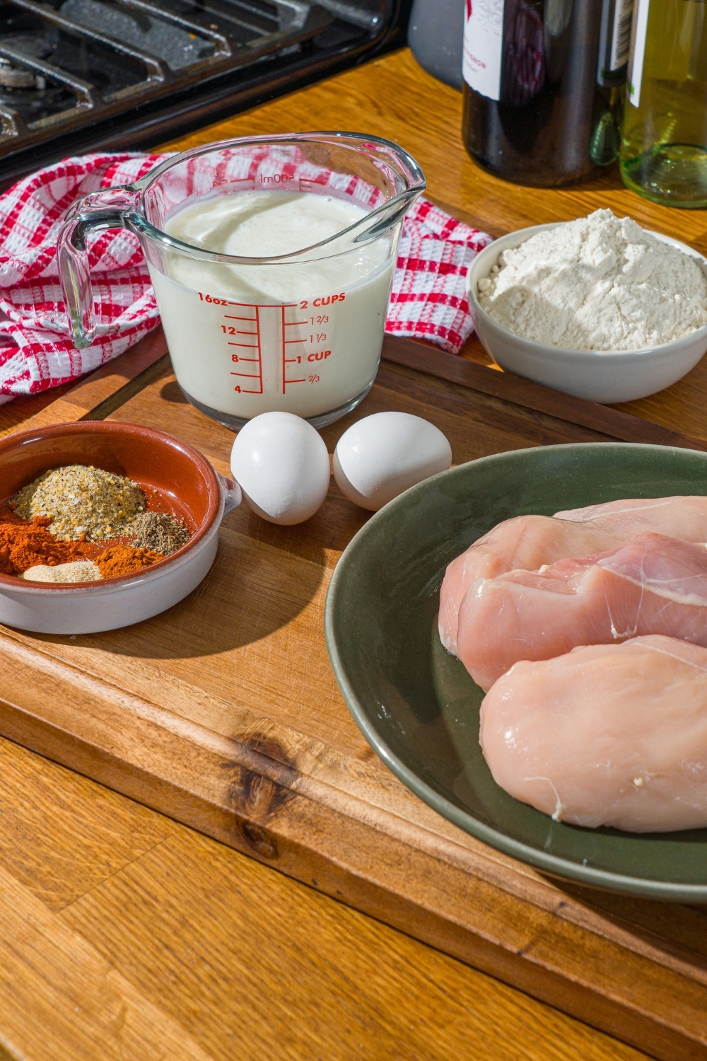 A wooden board with ingredients to make country fried chicken including chicken breast, buttermilk, flour, eggs, and seasonings. The board is on a wooden counter.