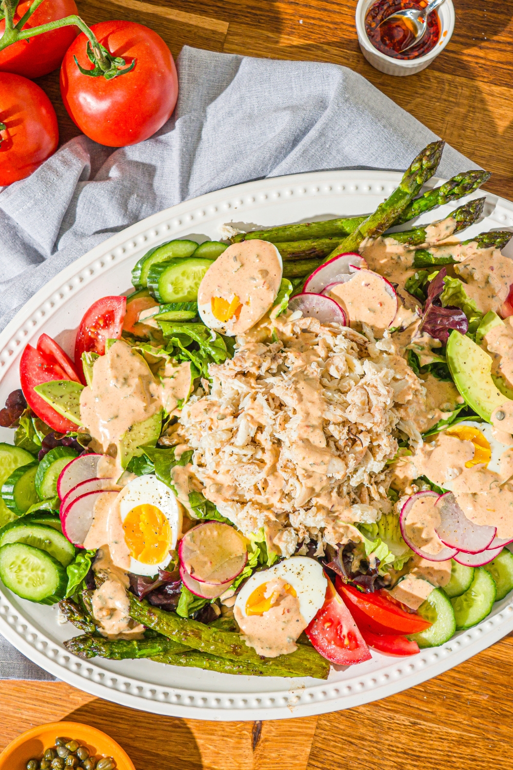 An oval platter with crab Louie salad including a bed of lettuce, hard boiled egg, asparagus, sliced vegetables, and lump crab drizzled with a crab Louie salad dressing. The platter is on a wooden counter with a blue cloth napkin and a bunch of tomatoes.