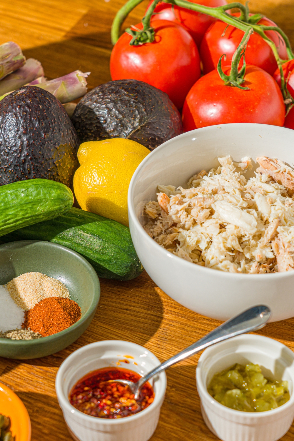A wooden counter with ingredients to make crab Louie salad including avocado, lemon, cucumber, lump crab, relish, and seasonings.