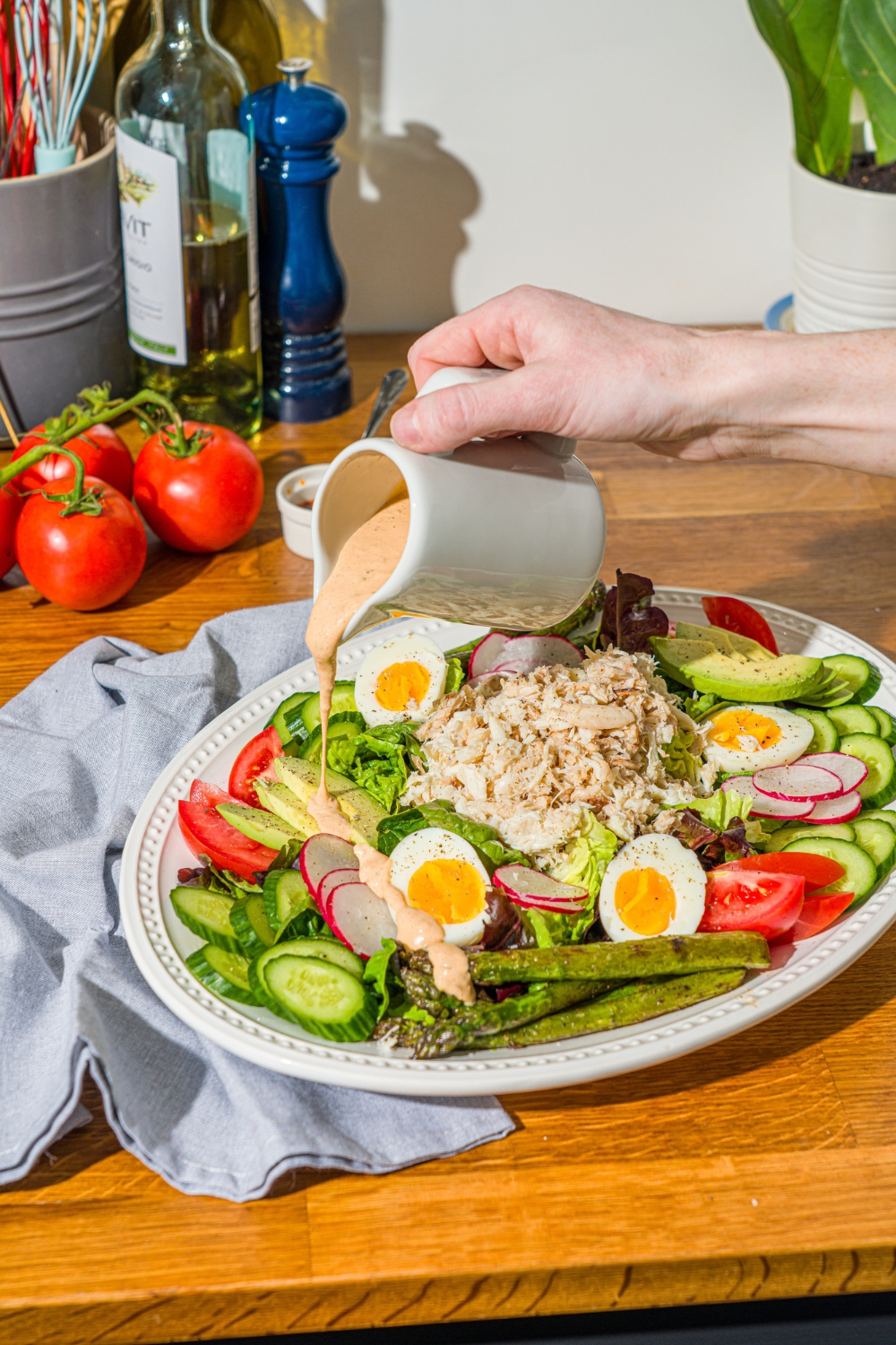An oval platter with crab Louie salad including a bed of lettuce, hard boiled egg, asparagus, sliced vegetables, and lump crab. A crab Louie salad dressing is being drizzled over the salad. The platter is on a wooden counter with a blue cloth napkin and a small bowl of capers.
