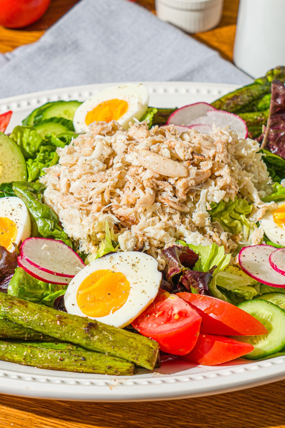 An oval platter with crab Louie salad including a bed of lettuce, hard boiled egg, asparagus, sliced vegetables, and lump crab. The platter is on a wooden counter with a blue cloth napkin.