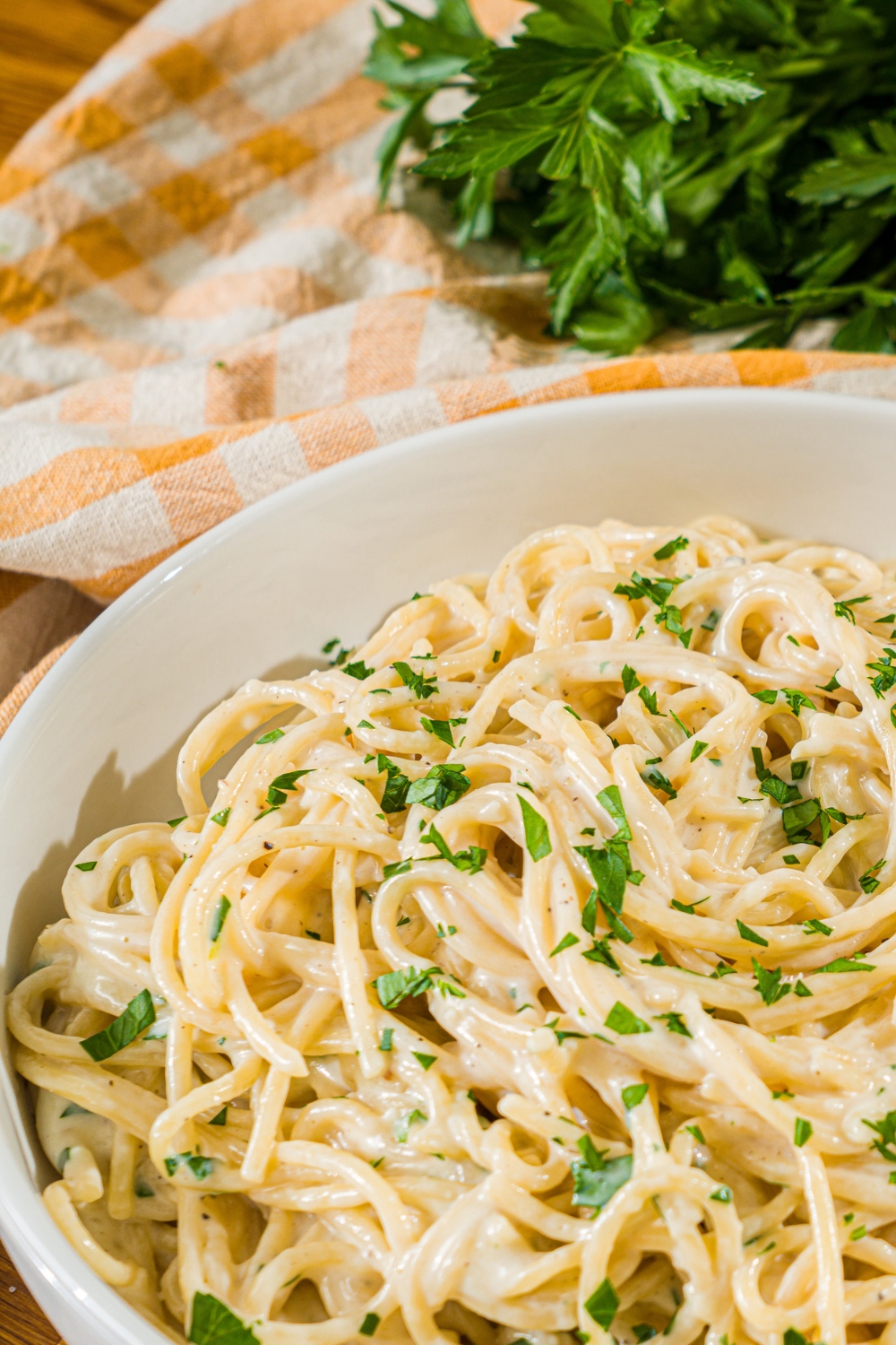 A white bowl with spaghetti tossed in a creamy garlic parmesan sauce garnished with fresh parsley. The bowl is on a wooden counter with a yellow checkered napkin and bunch of fresh parsley.