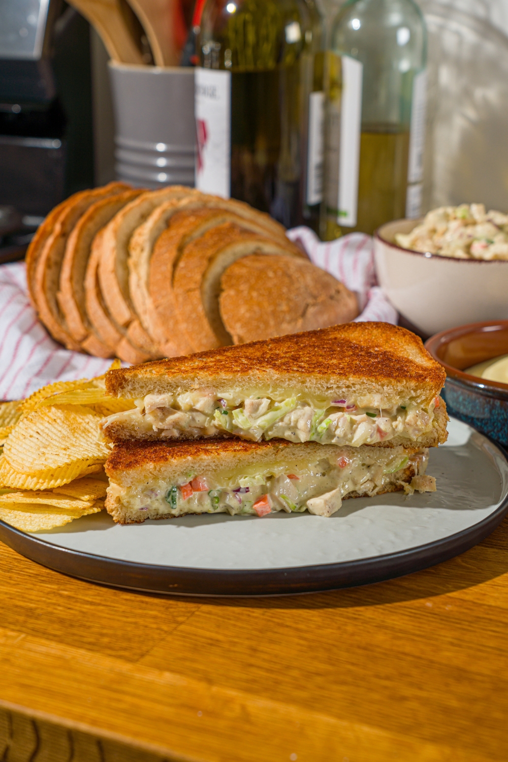 A white plate with a chicken melt sandwich sliced in half and stacked on one another. The plate is served with chips. The plate is on a wooden counter with a basket of sliced bread.