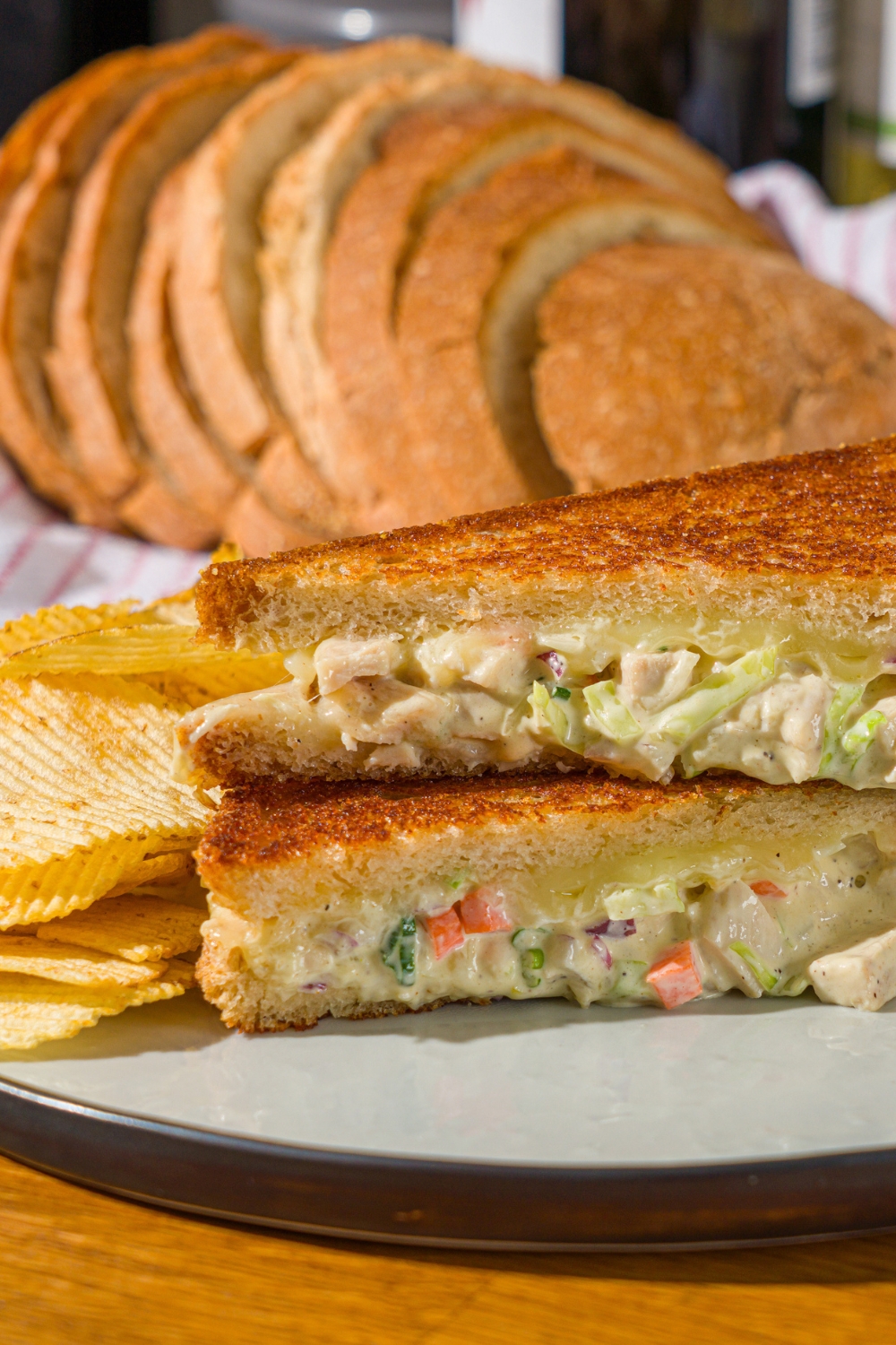 A white plate with a chicken melt sandwich sliced in half and stacked on one another. The plate is served with chips. The plate is on a wooden counter with a basket of sliced bread.