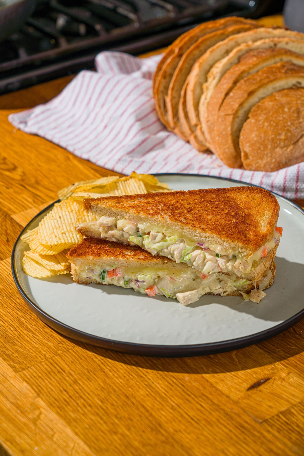 A white plate with a chicken melt sandwich sliced in half and stacked on one another. The plate is served with chips. The plate is on a wooden counter with a basket of sliced bread.