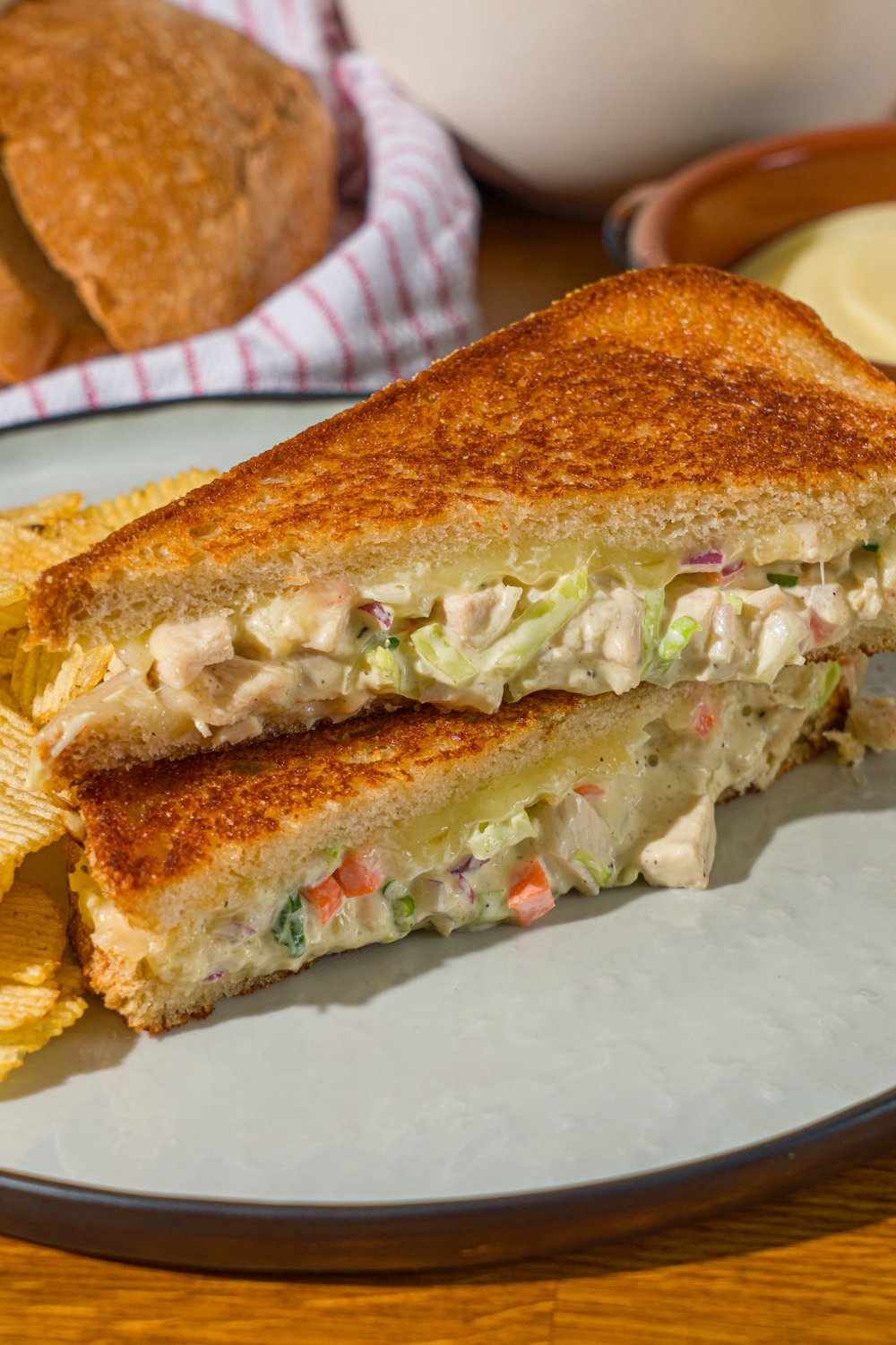 A white plate with a chicken melt sandwich sliced in half and stacked on one another. The plate is served with chips. The plate is on a wooden counter with a basket of sliced bread.