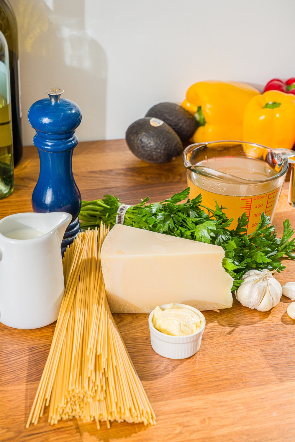 A wooden counter with ingredients to make creamy garlic parmesan sauce including parmesan cheese, milk, butter, garlic, chicken broth, spaghetti, and fresh parsley.