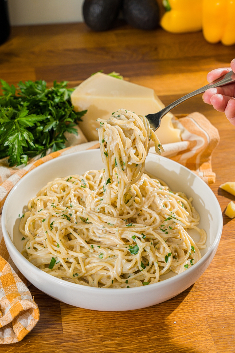 A white bowl with spaghetti tossed in a creamy garlic parmesan sauce garnished with fresh parsley. A fork is taking a bite of pasta from the bowl. The bowl is on a wooden counter with a yellow checkered napkin.