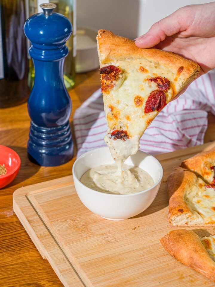 A hand dipping a slice of white pizza with roasted peppers into a bowl of garlic white pizza sauce. The bowl is on a wooden board with additional slices of pizza.