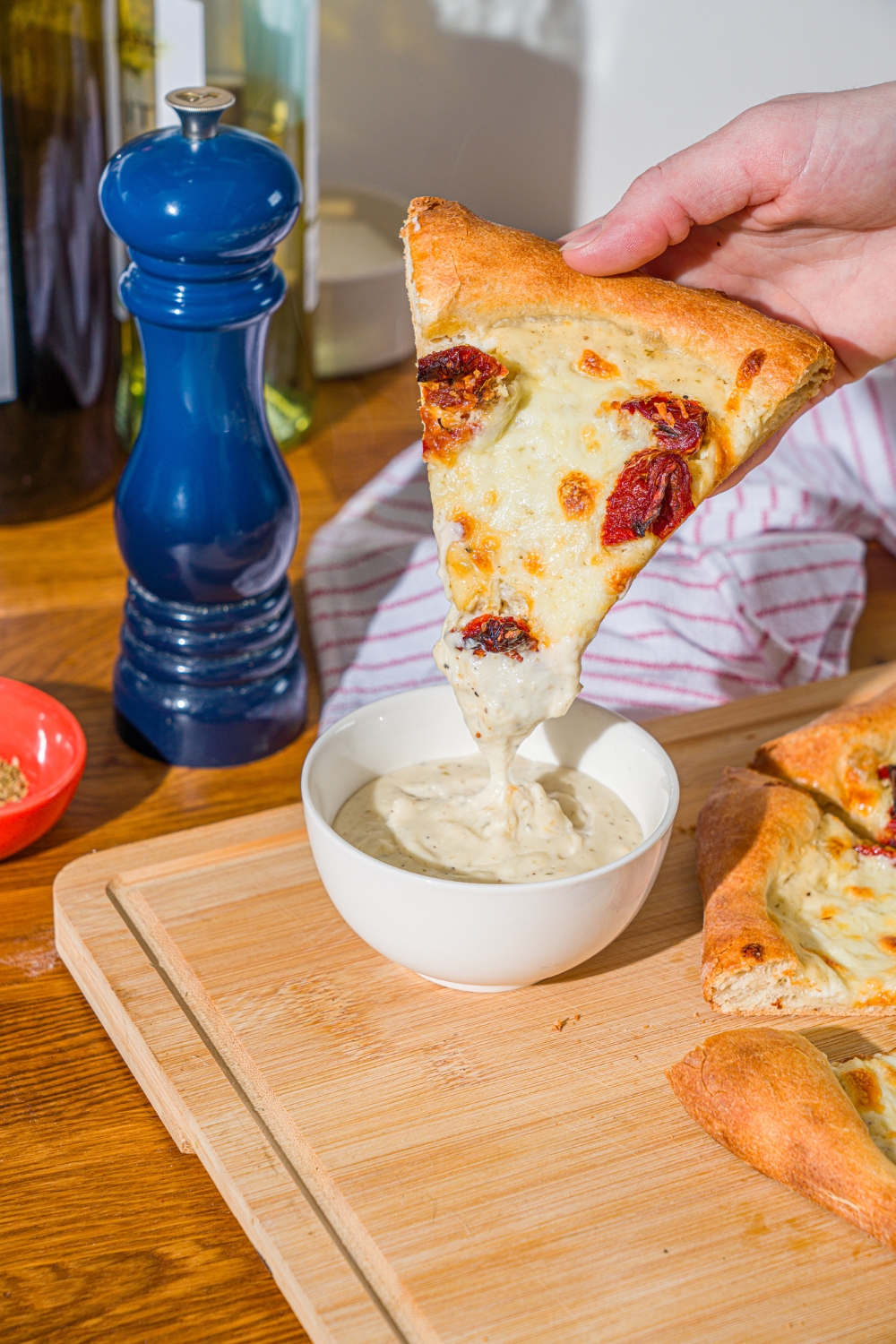 A hand dipping a slice of white pizza with roasted peppers into a bowl of garlic white pizza sauce. The bowl is on a wooden board with additional slices of pizza.