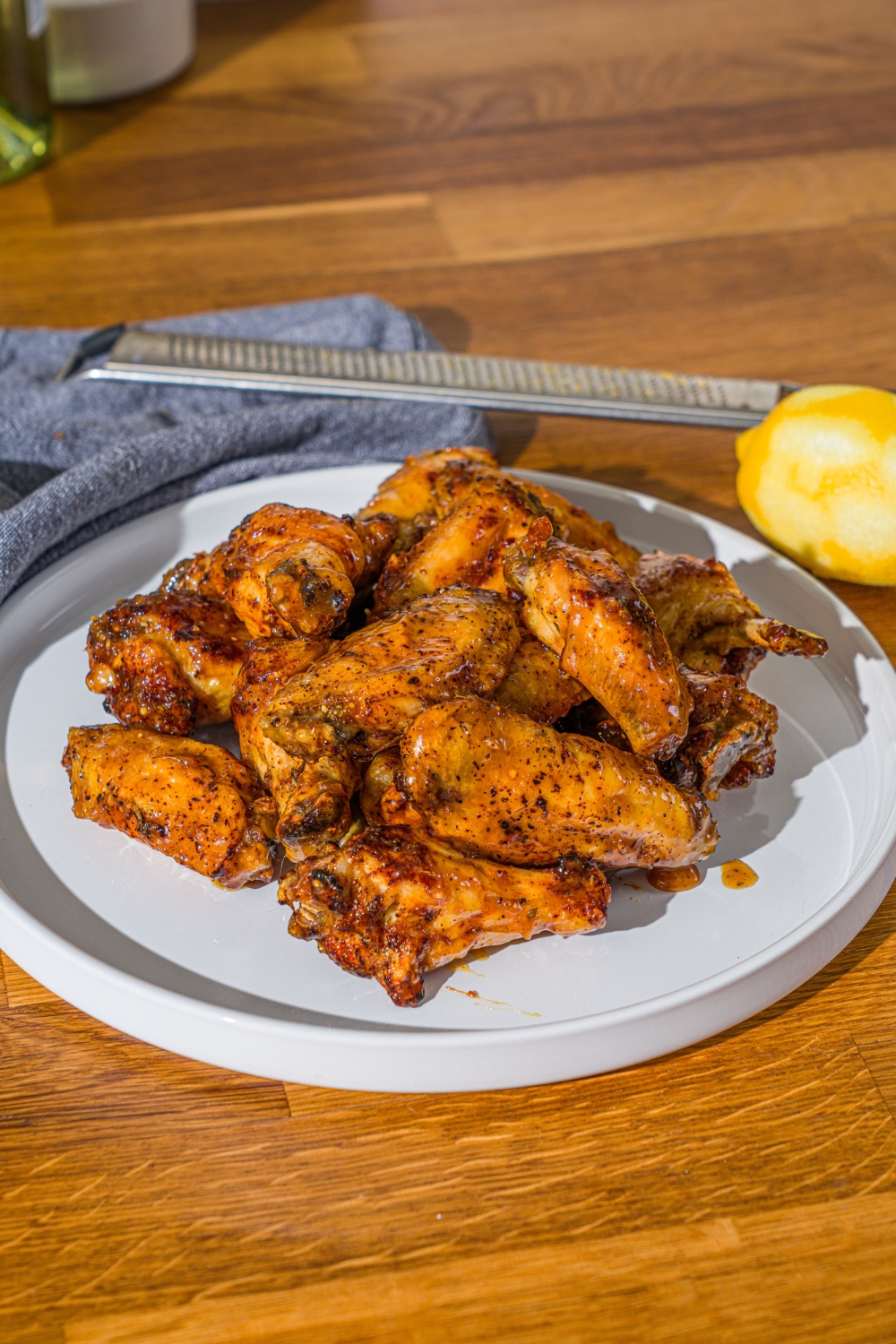 A white plate with a pile of crispy honey lemon pepper wings. The plate is on a wooden counter with a blue cloth napkin and a zested lemon.