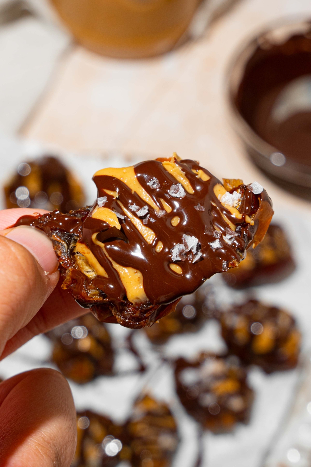A close up of a hand holding a date stuffed with peanut butter and topped with chocolate and sea salt. There is a plate of peanut butter dates in the background.