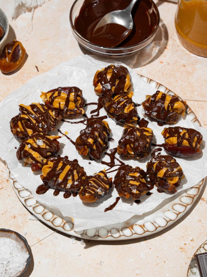 A plate lined with parchment paper with dates stuffed with peanut butter and drizzled with chocolate and sea salt. The plate is on a tan counter with a small plate of sea salt and bowl of melted chocolate.