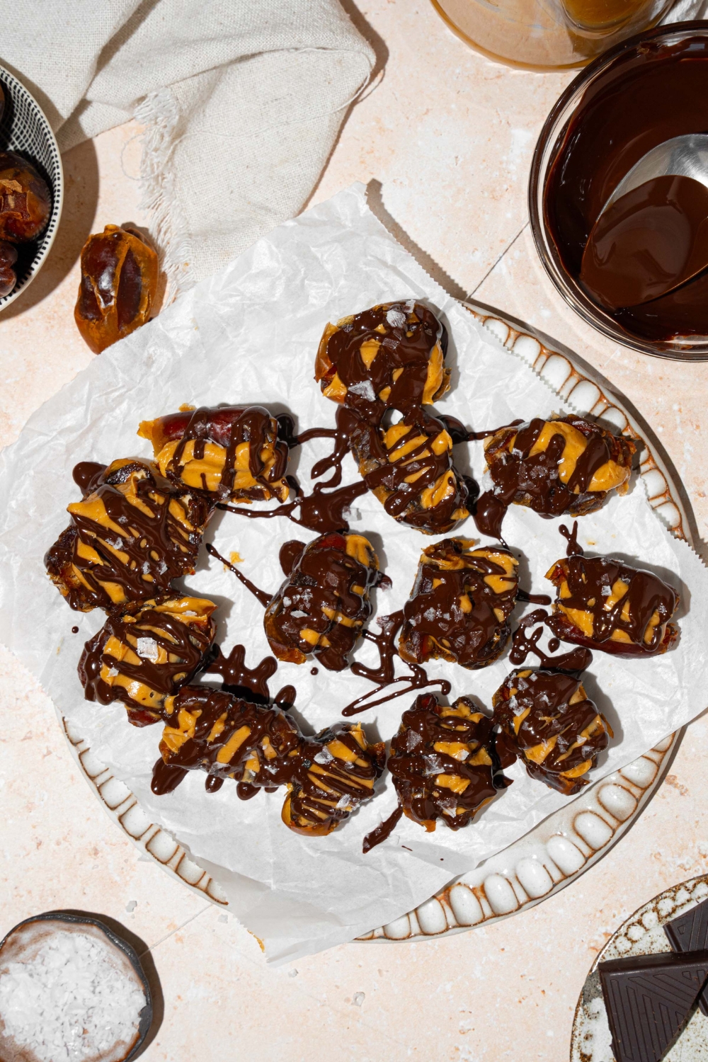 A plate lined with parchment paper with dates stuffed with peanut butter and drizzled with chocolate and sea salt. The plate is on a tan counter with a small plate of sea salt and bowl of melted chocolate.