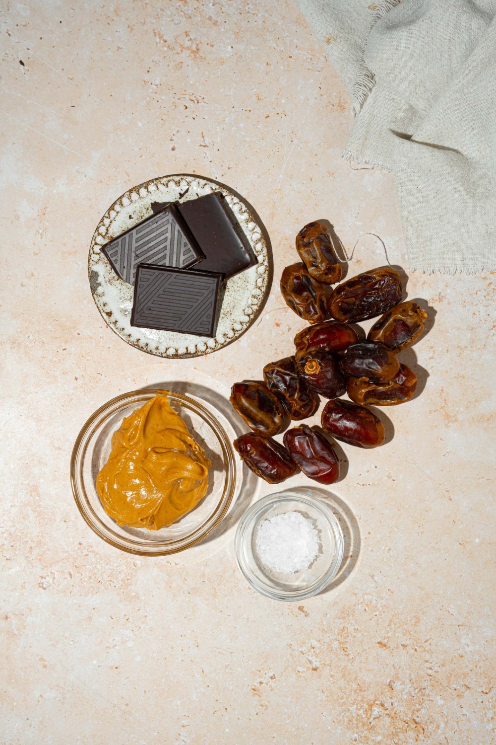 A tan counter with ingredients to make dates with peanut butter including medjool dates, peanut butter, dark chocolate, and sea salt.