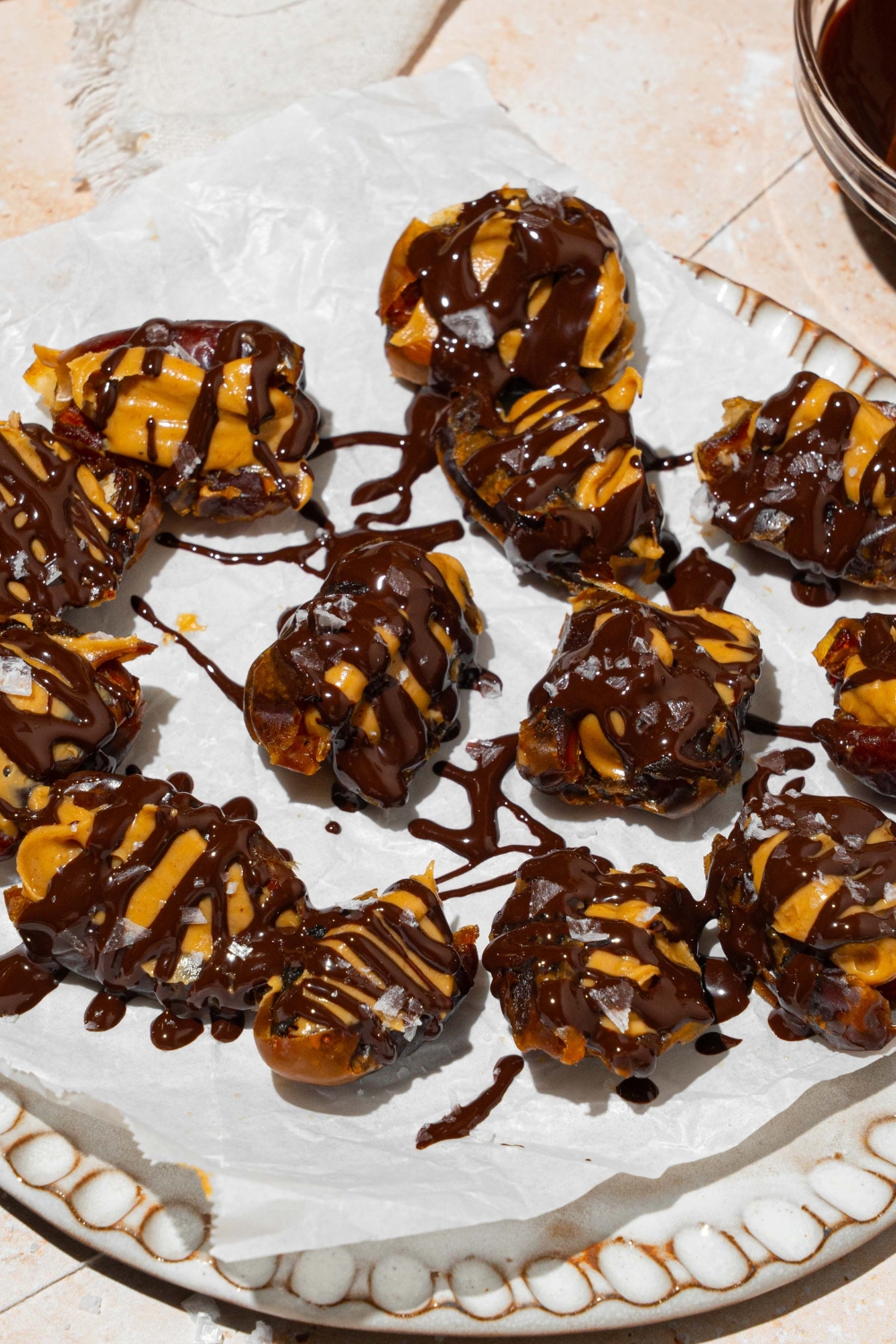 A plate lined with parchment paper with dates stuffed with peanut butter and drizzled with chocolate and sea salt. The plate is on a tan counter.