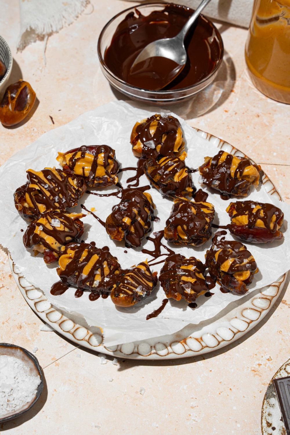 A plate lined with parchment paper with dates stuffed with peanut butter and drizzled with chocolate and sea salt. The plate is on a tan counter with a small plate of sea salt and bowl of melted chocolate.