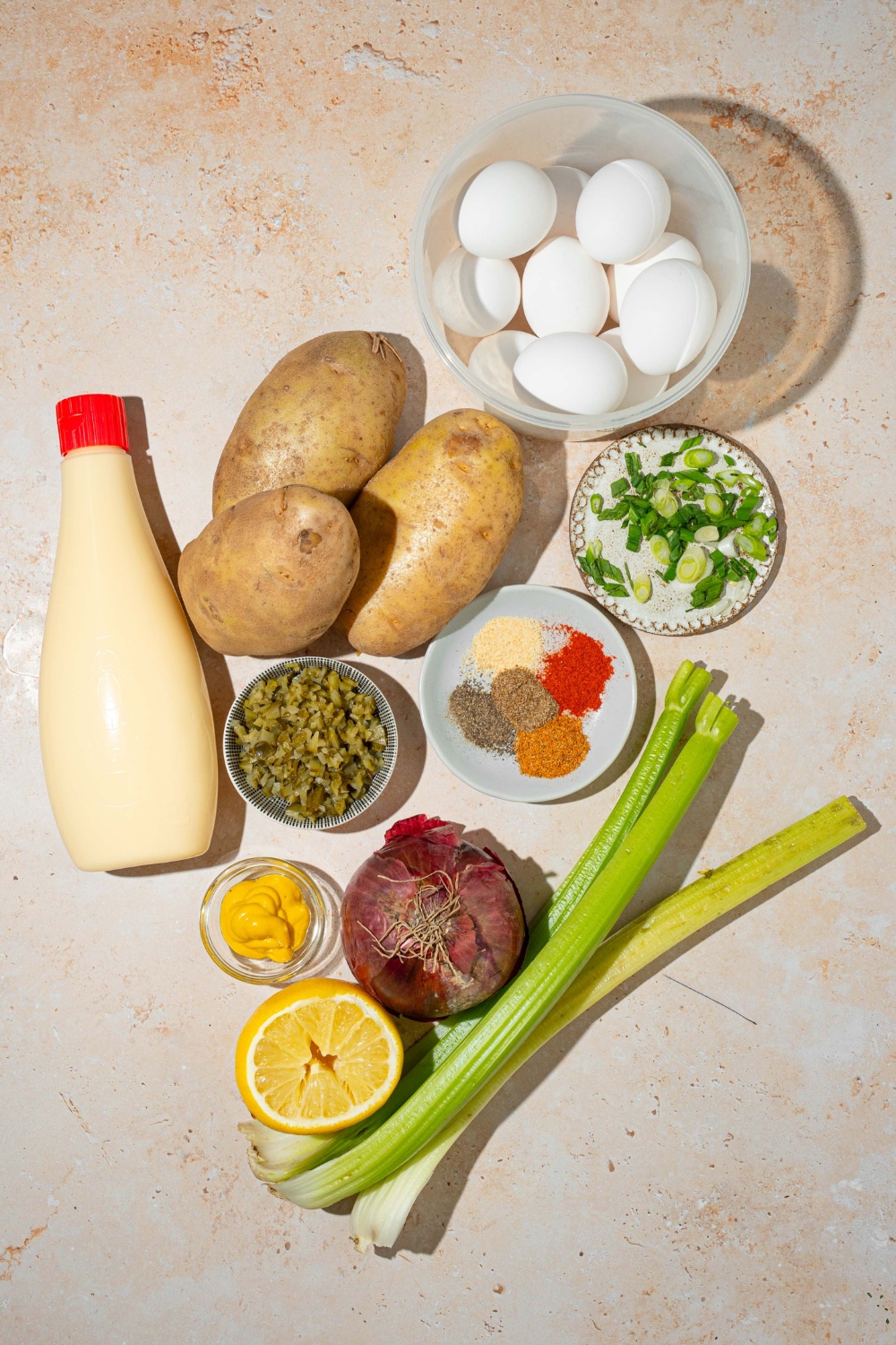 A tan counter with ingredients to make deviled egg potato salad including eggs, potatoes, celery, red onion, lemon, mustard, relish, mayonnaise, and seasonings.