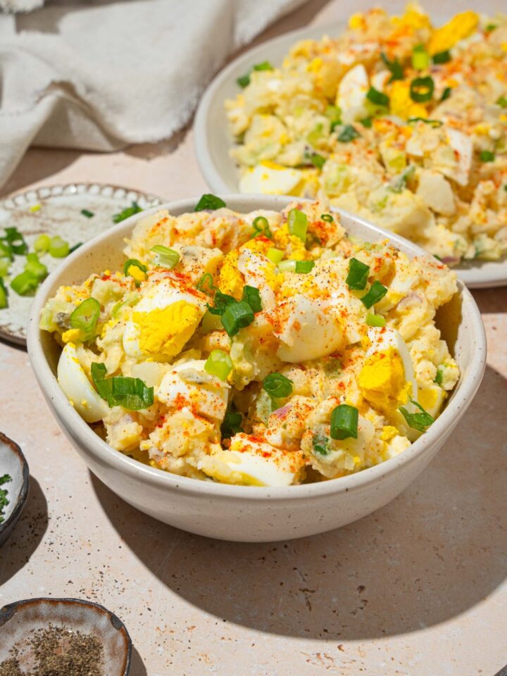 A white bowl with deviled egg potato salad garnished with sliced green egg and paprika. The bowl is on a tan counter with an additional plate of potato salad.