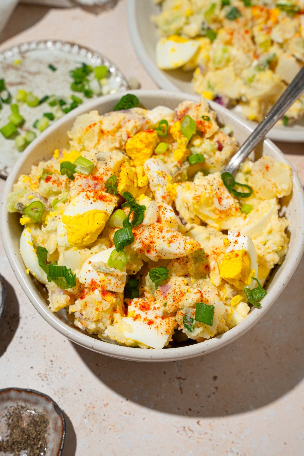A white bowl with deviled egg potato salad garnished with sliced green egg and paprika. There is a fork taking a bite from the bowl. The bowl is on a tan counter with an additional plate of potato salad.