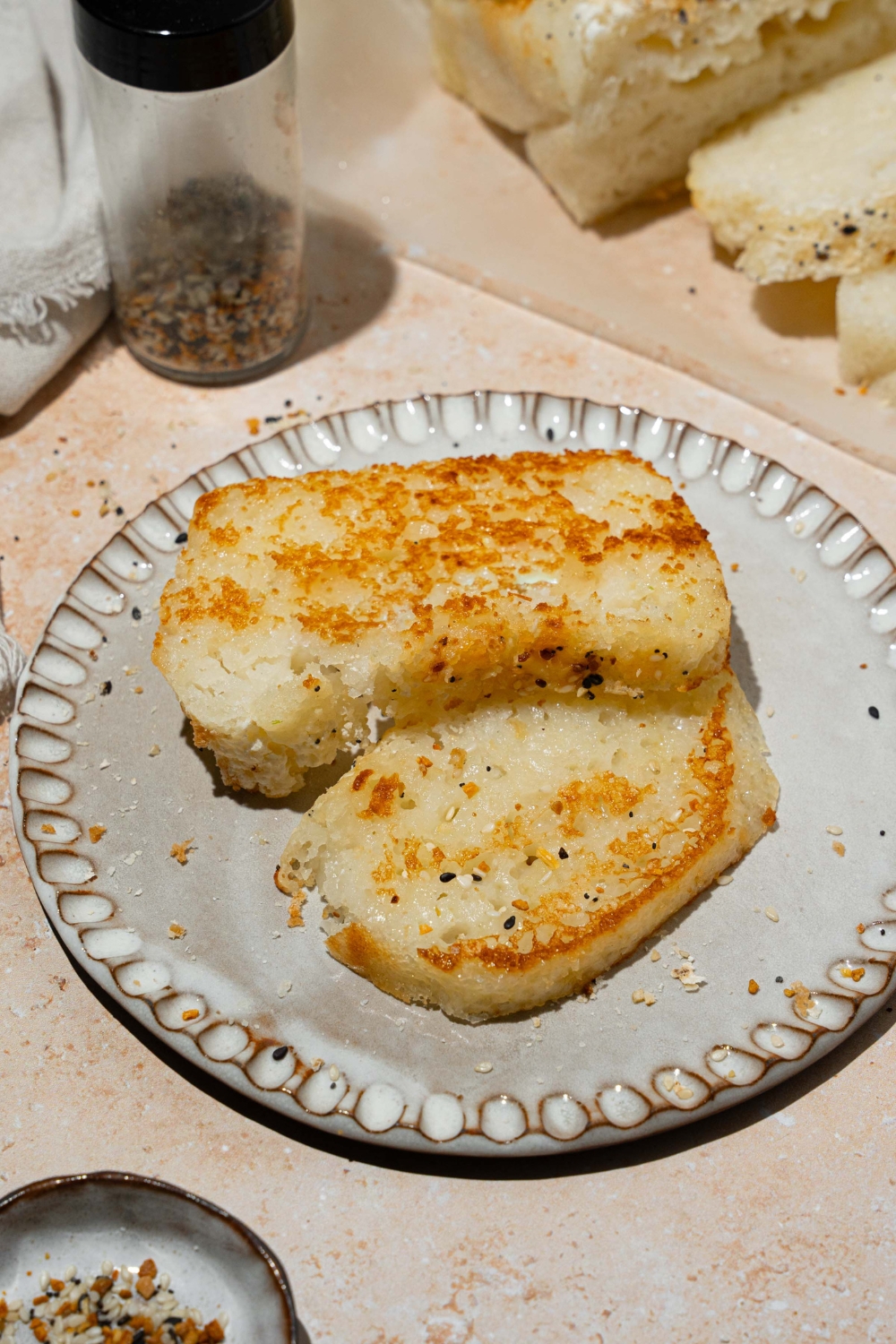 A white plate with two slices of blender rice bread. The plate is on a tan counter with a loaf of sliced bread topped with everything bagel seeds.