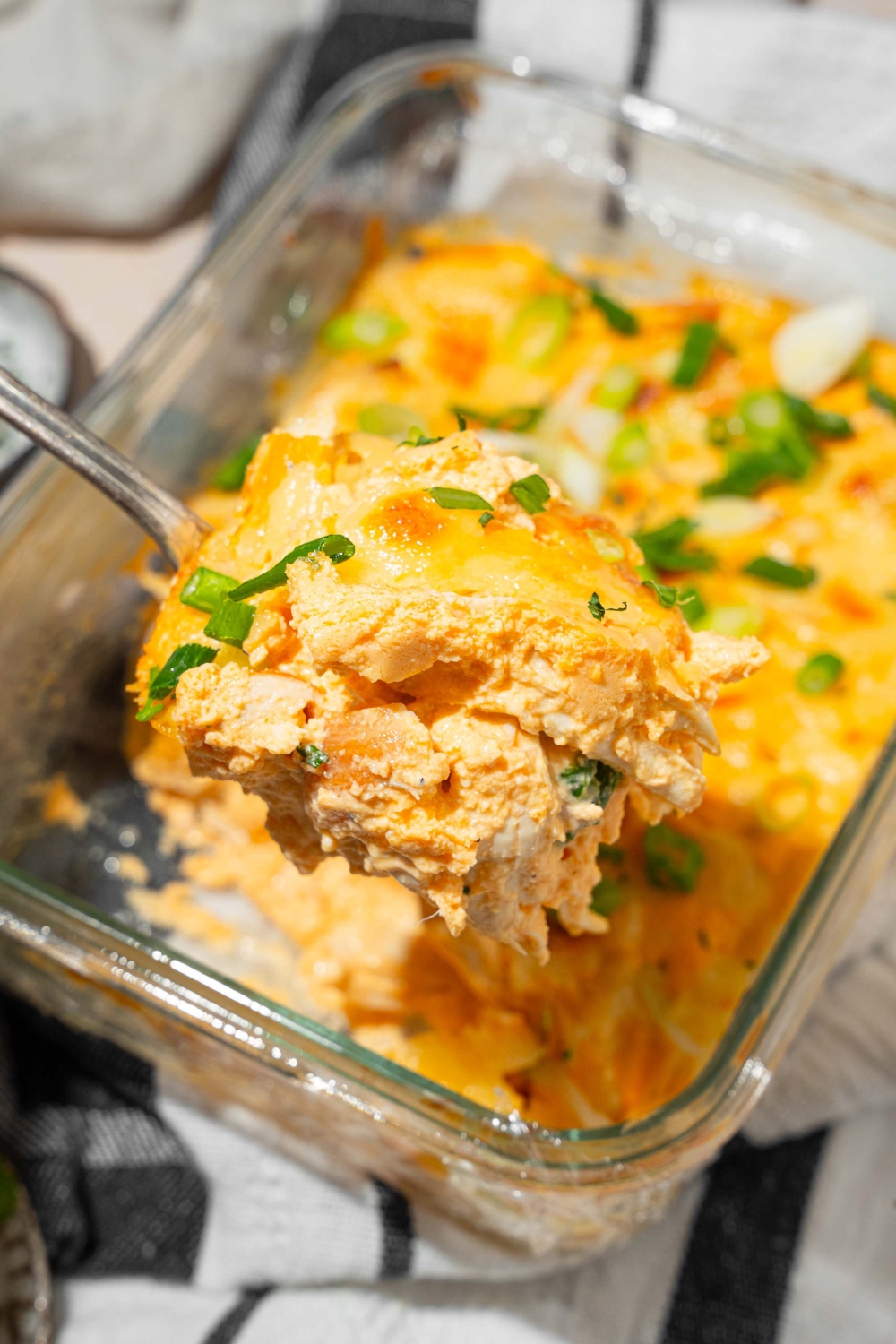 A close up of a spoon with a bite of cottage cheese Buffalo chicken dip garnished with green onion. There is a baking dish of dip blurred in the background.